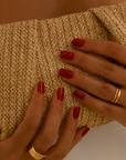 Close-up of hands holding a textured beige clutch with red nail polish and gold rings.