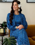 A woman wearing a blue and black patterned long-sleeve dress sits on a mustard yellow armchair in a living room setting, with a side table, potted plants, and framed wall art in the background.