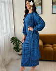 A woman models a long-sleeved blue maxi dress with a white geometric tribal pattern in a brightly lit room with a brown velvet sofa and a potted plant.