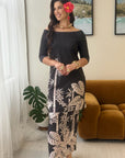 A woman models an off-the-shoulder maxi dress with a black base and a beige tropical leaf and bamboo print. She is standing in a modern living room setting with a brown velvet sofa, framed landscape art, and a potted plant.