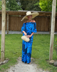 Woman in a blue dress and wide-brimmed hat standing under a wooden archway with greenery around.