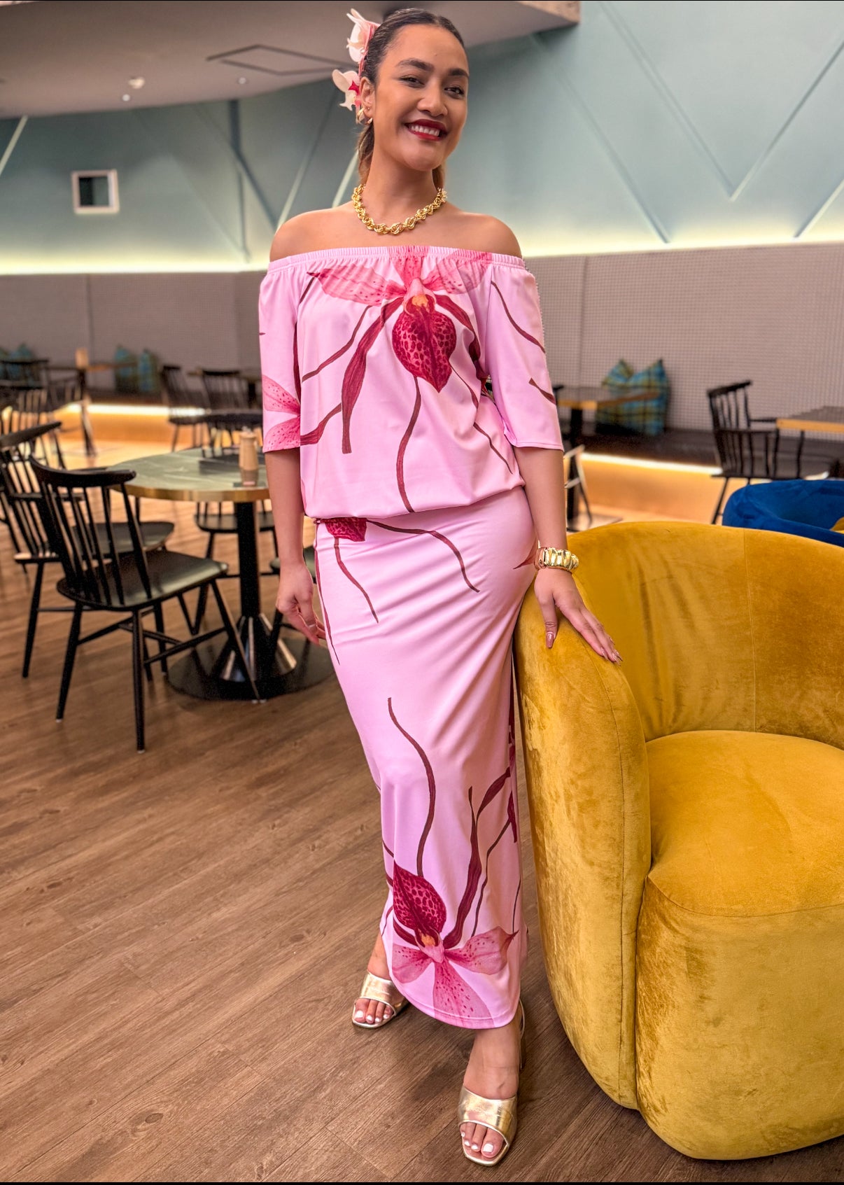 Woman in a pink floral off-shoulder top and skirt standing in a modern interior setting.