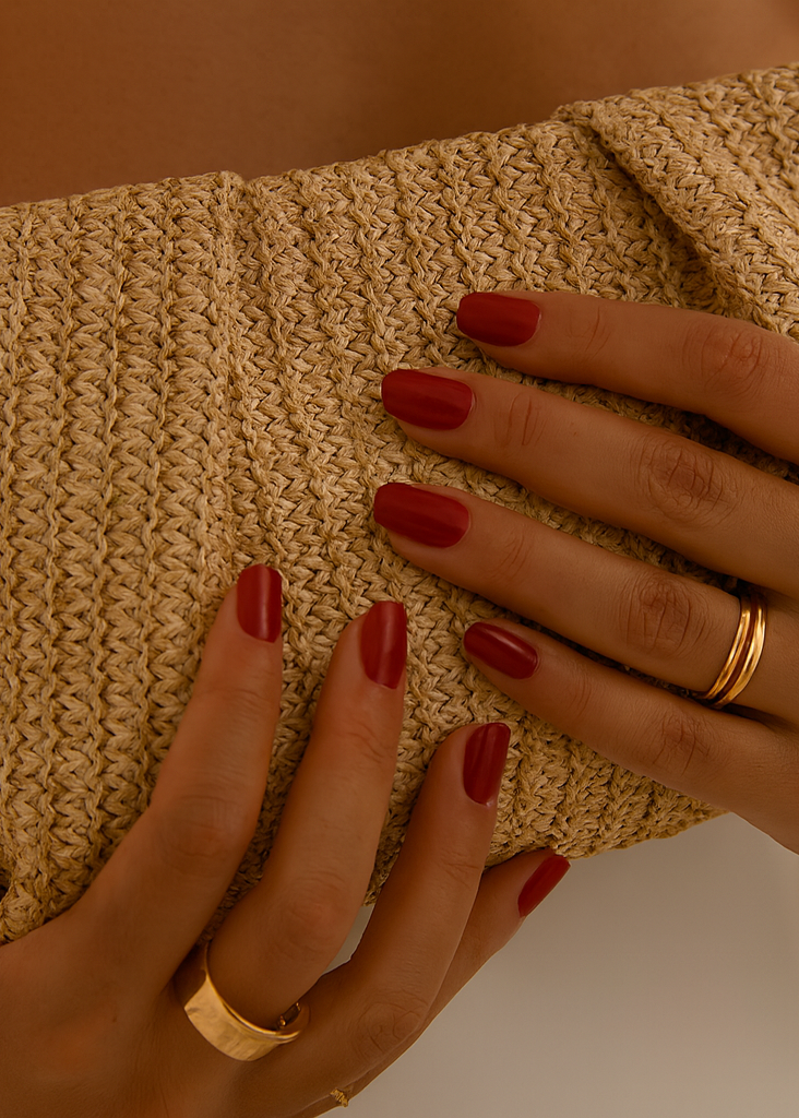 Close-up of hands holding a textured beige clutch with red nail polish and gold rings.