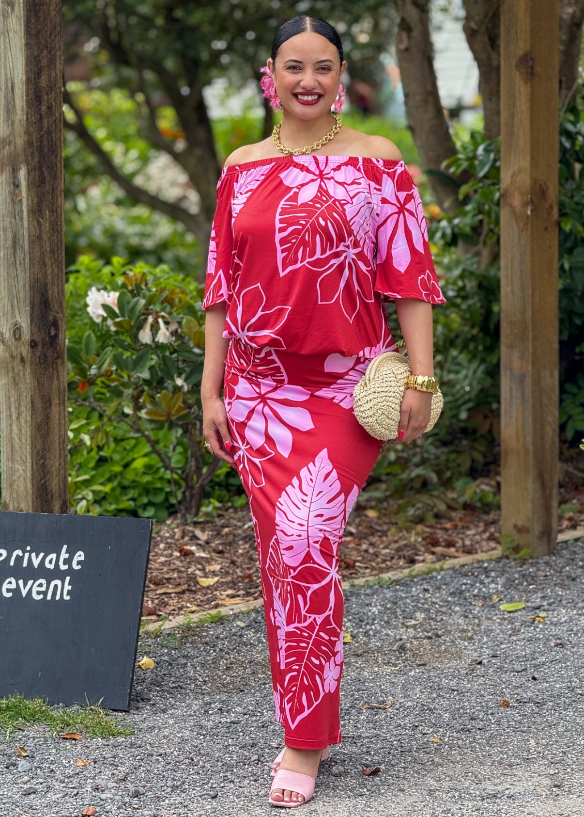 Woman in a red and pink floral outfit standing outdoors with greenery in the background.