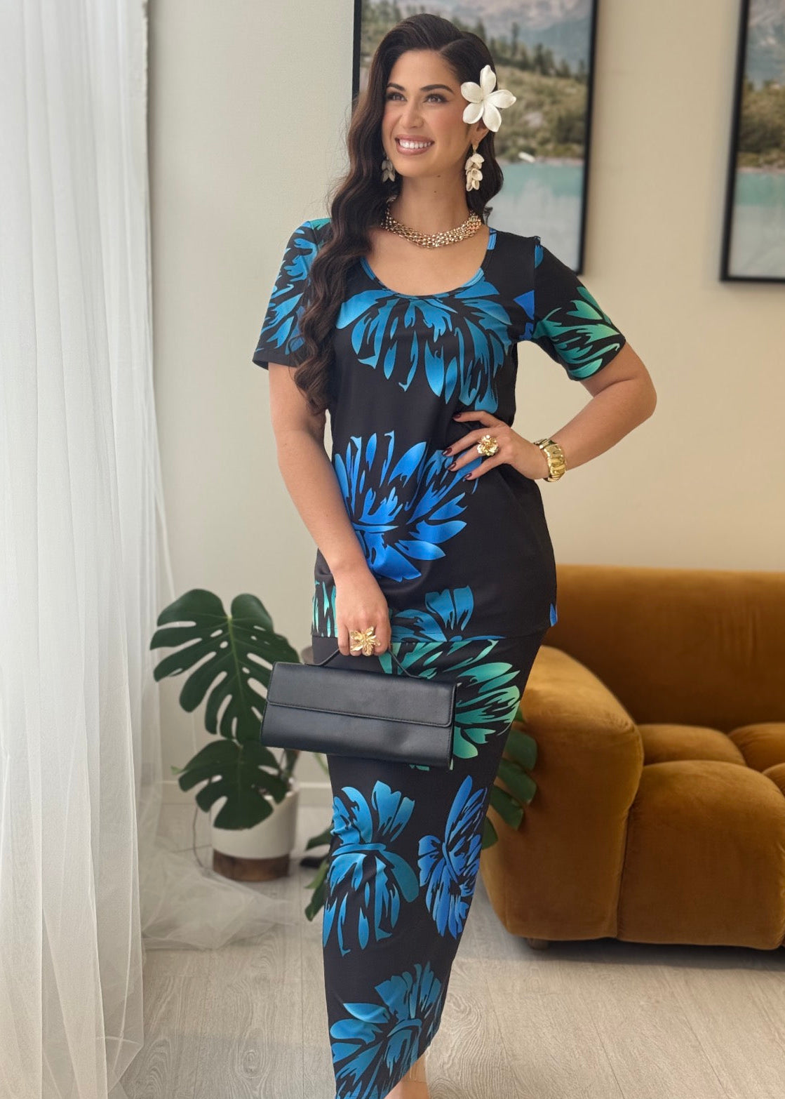 A woman models a short-sleeve midi dress with a black base and a vibrant blue and green tropical floral pattern. She is standing in a living room setting with a brown velvet sofa, a potted plant, and a white curtain in the background.