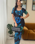 A woman models a short-sleeve midi dress with a black base and a vibrant blue and green tropical floral pattern. She is standing in a living room setting with a brown velvet sofa, a potted plant, and a white curtain in the background.