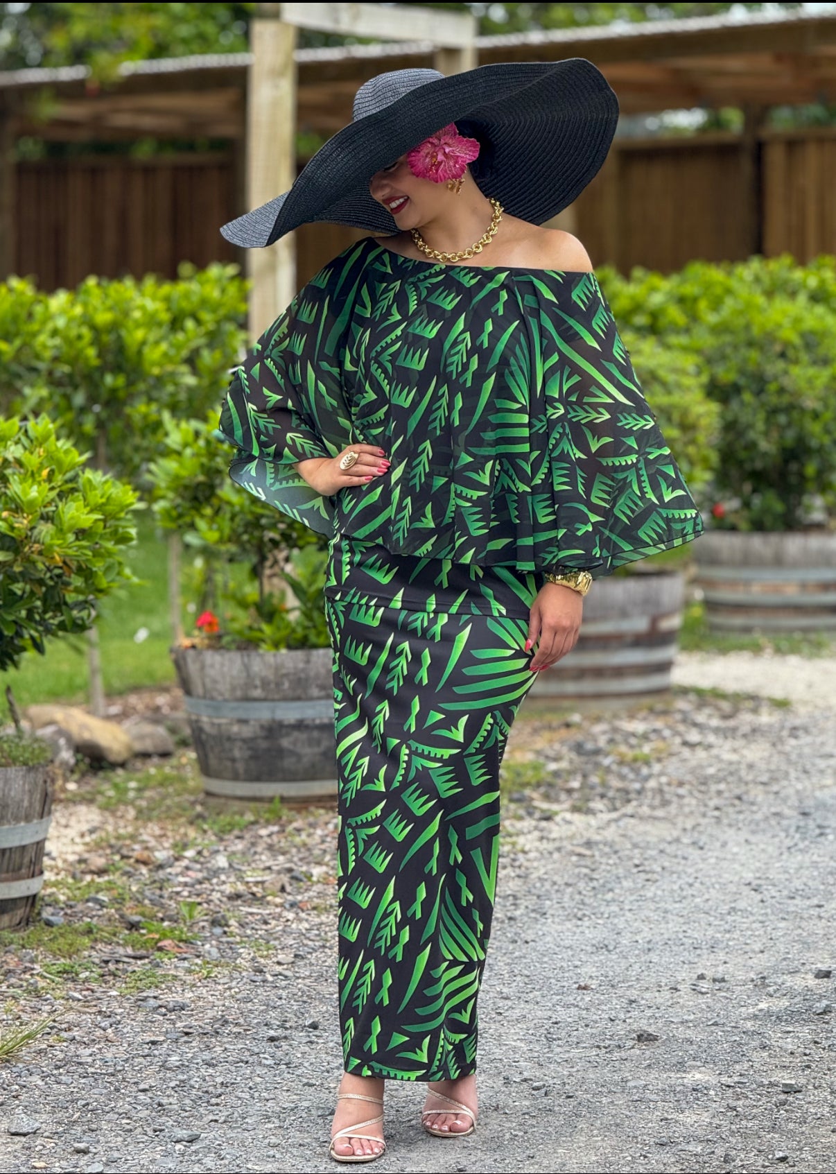 Woman in a green and black leaf-patterned outfit with a large black hat and pink flower, standing outdoors.