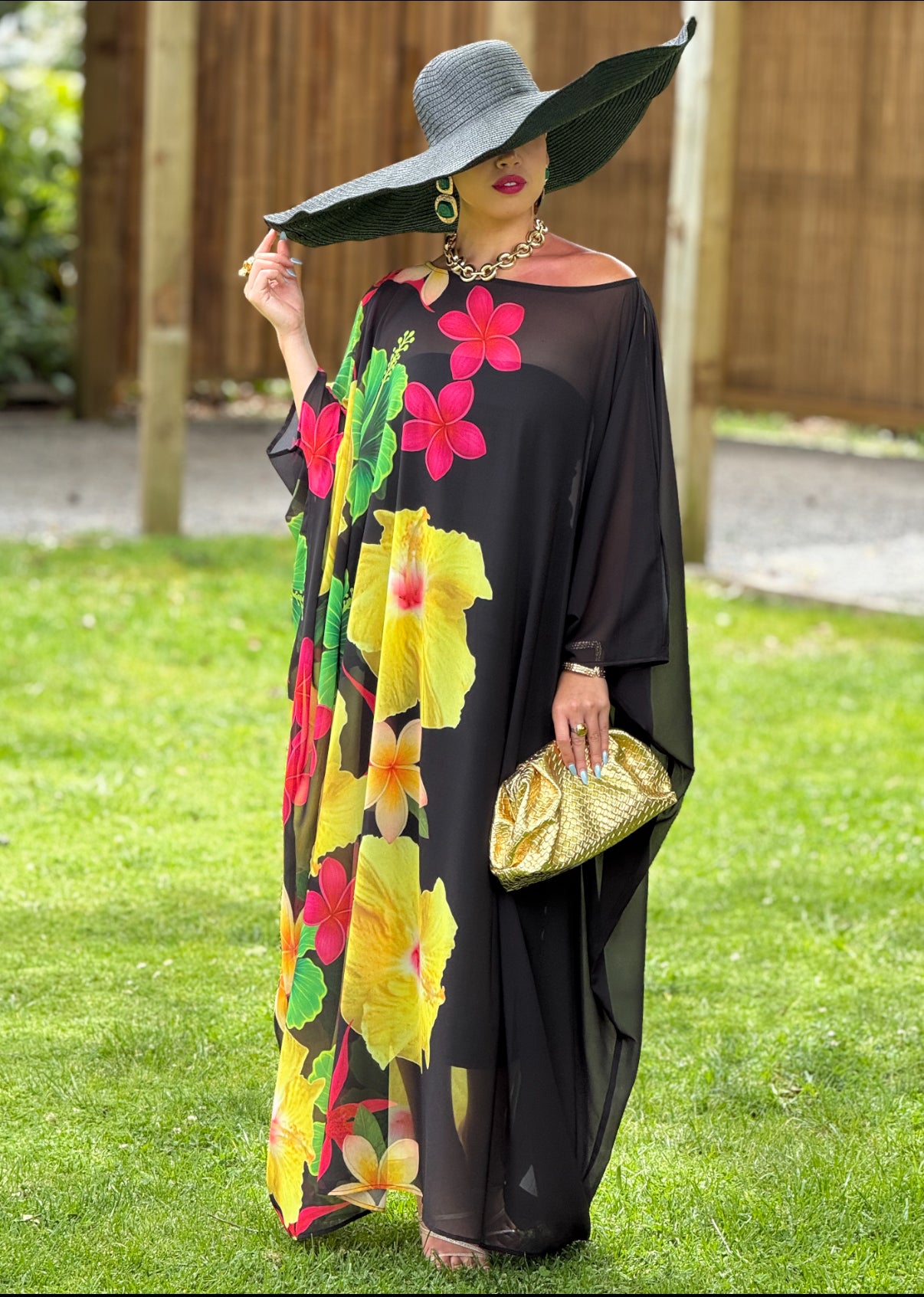 Woman in a colorful floral dress and large hat standing on grass.
