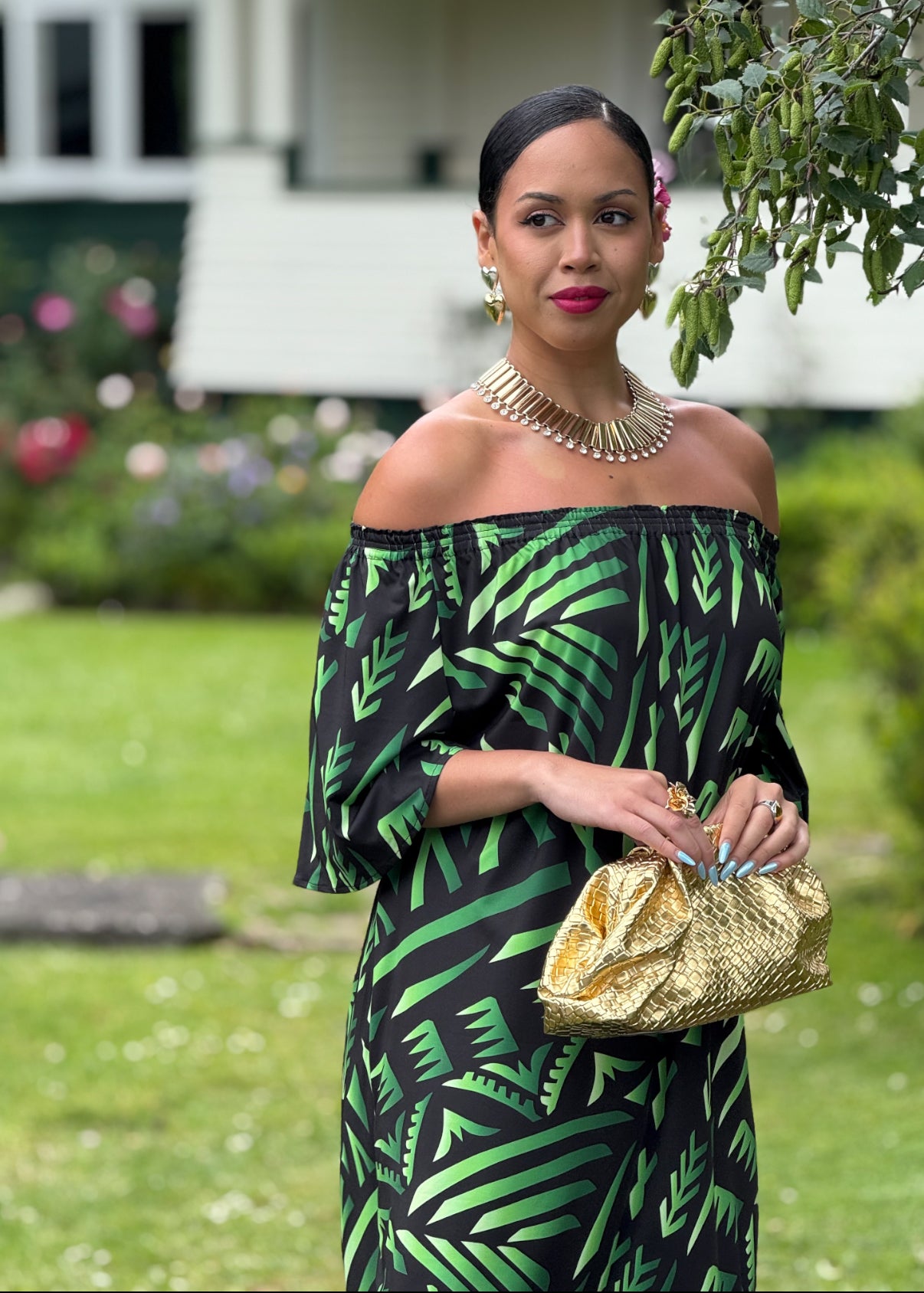 Woman in a green and black patterned dress holding a gold clutch in an outdoor setting.