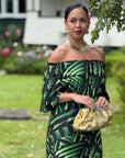 Woman in a green and black patterned dress holding a gold clutch in an outdoor setting.