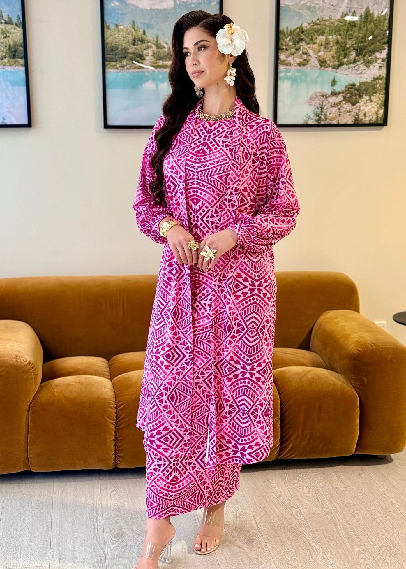 A woman models a long-sleeved magenta pink kaftan dress with a white geometric pattern, standing in front of a brown velvet sofa and framed landscape prints on a beige wall.