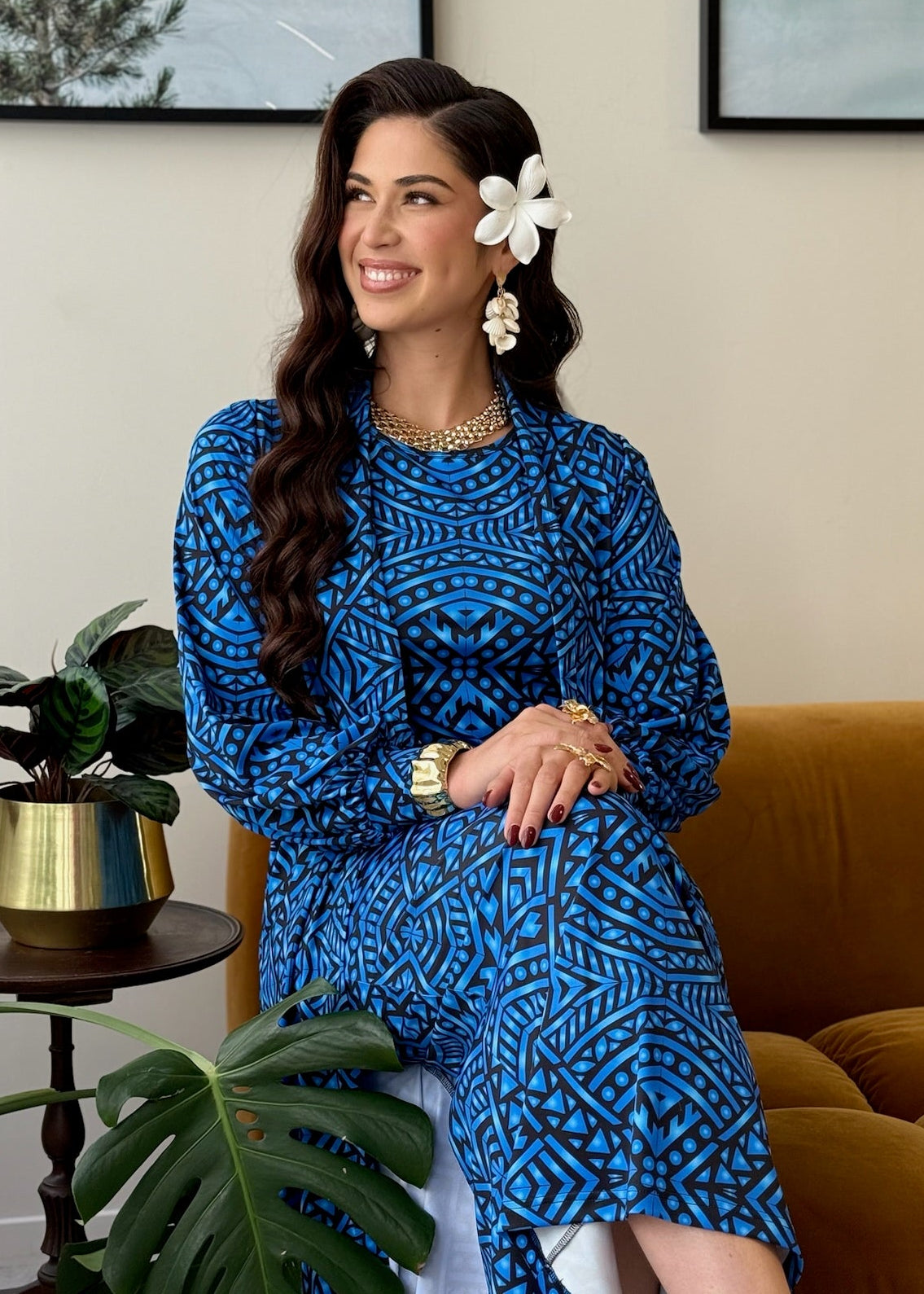 A woman wearing a blue and black patterned long-sleeve dress sits on a mustard yellow armchair in a living room setting, with a side table, potted plants, and framed wall art in the background.