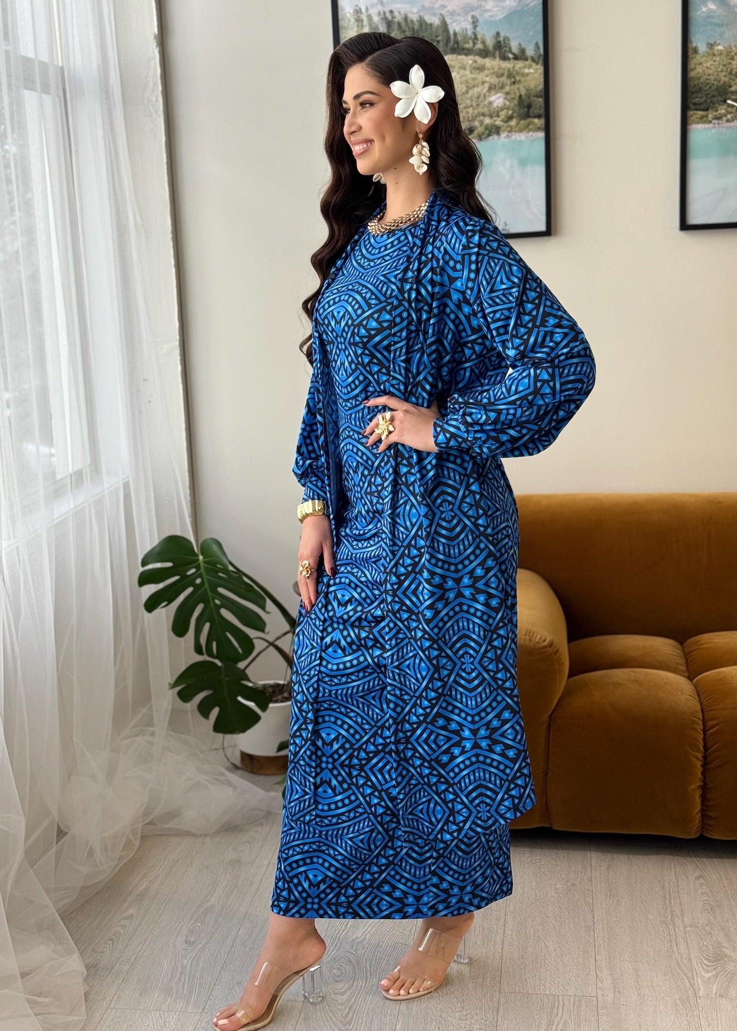 A woman models a long-sleeved blue maxi dress with a white geometric tribal pattern in a brightly lit room with a brown velvet sofa and a potted plant.