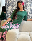 A woman wearing a green three-quarter sleeve dress with pink tropical floral prints sits on a cream boucle sofa in a modern living room setting with floral wallpaper.