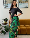 A woman models a black maxi dress with a green tropical leaf print on the skirt, standing in a living room setting with a brown velvet sofa and framed landscape art in the background.