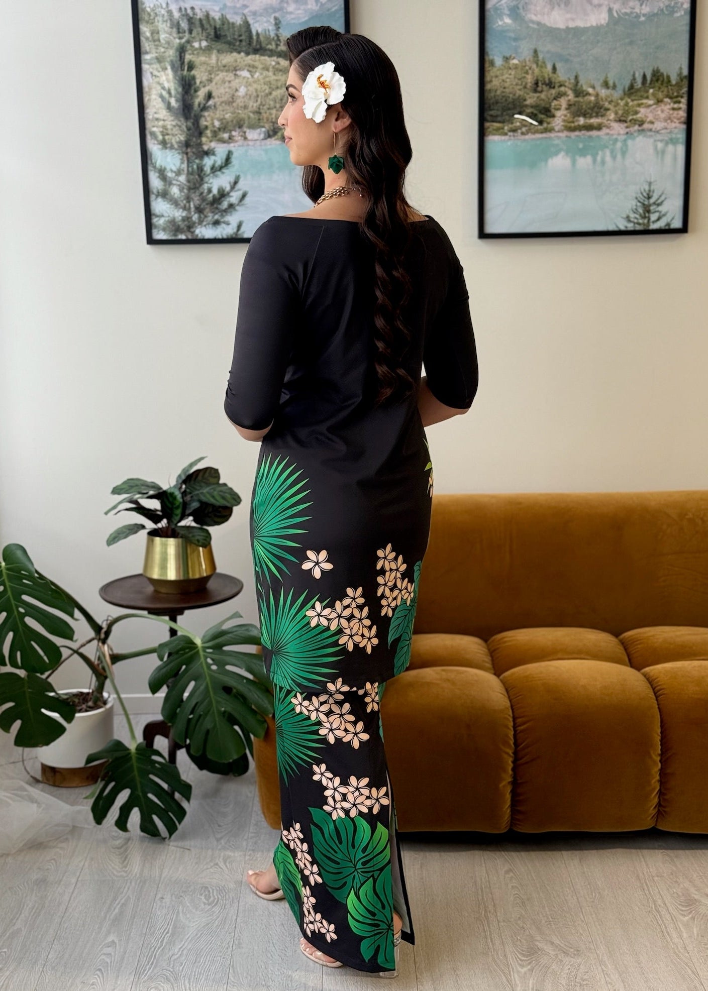 A woman stands with her back to the camera wearing a black long-sleeve maxi dress with a green palm leaf and beige floral print down the side. She is in a living room setting with a mustard yellow sofa, framed landscape pictures on the wall, and potted plants.