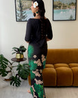 A woman stands with her back to the camera wearing a black long-sleeve maxi dress with a green palm leaf and beige floral print down the side. She is in a living room setting with a mustard yellow sofa, framed landscape pictures on the wall, and potted plants.
