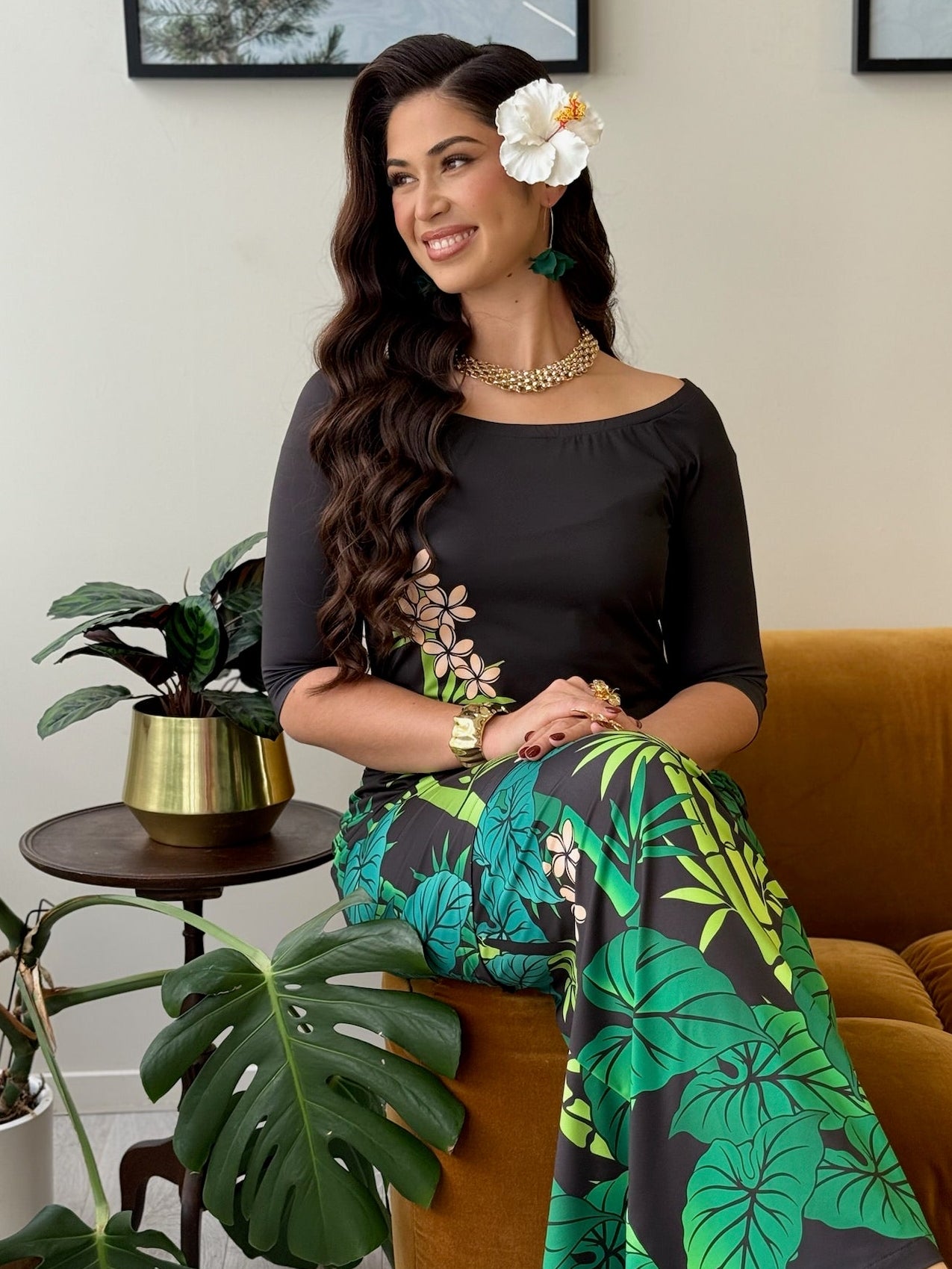A woman sits on a mustard-brown velvet sofa wearing a black off-the-shoulder top and matching black skirt with a green tropical leaf print. She has a white flower in her hair, a gold necklace, and is posed in a living room setting with potted plants and framed wall art.