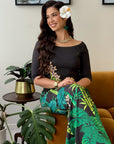 A woman sits on a mustard-brown velvet sofa wearing a black off-the-shoulder top and matching black skirt with a green tropical leaf print. She has a white flower in her hair, a gold necklace, and is posed in a living room setting with potted plants and framed wall art.