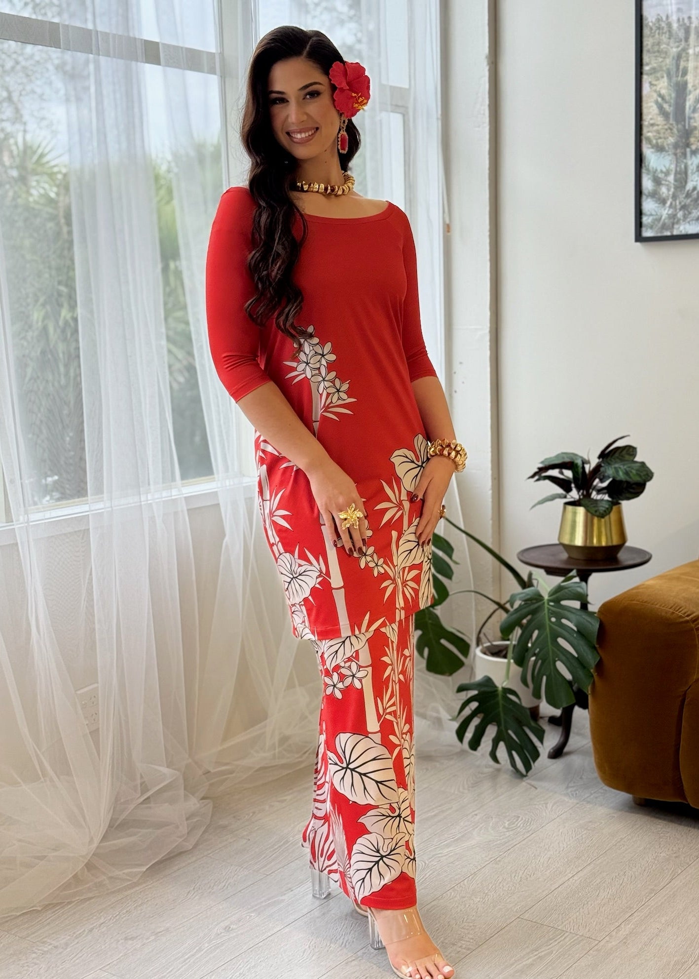 A woman models a red three-quarter sleeve maxi dress with a white tropical leaf print, standing in a bright, modern room with sheer white curtains, a brown armchair, and a potted plant.