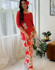 A woman models a red three-quarter sleeve maxi dress with a white tropical leaf print, standing in a bright, modern room with sheer white curtains, a brown armchair, and a potted plant.
