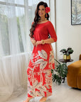 A woman models a red maxi dress with a beige palm leaf pattern in a bright, modern room with sheer curtains, a plant, and a mustard-colored armchair.