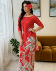 A woman models a red three-quarter sleeve kurta set with a white and gray tropical leaf print, styled with gold jewelry and a red flower hair accessory in a bright living room setting.