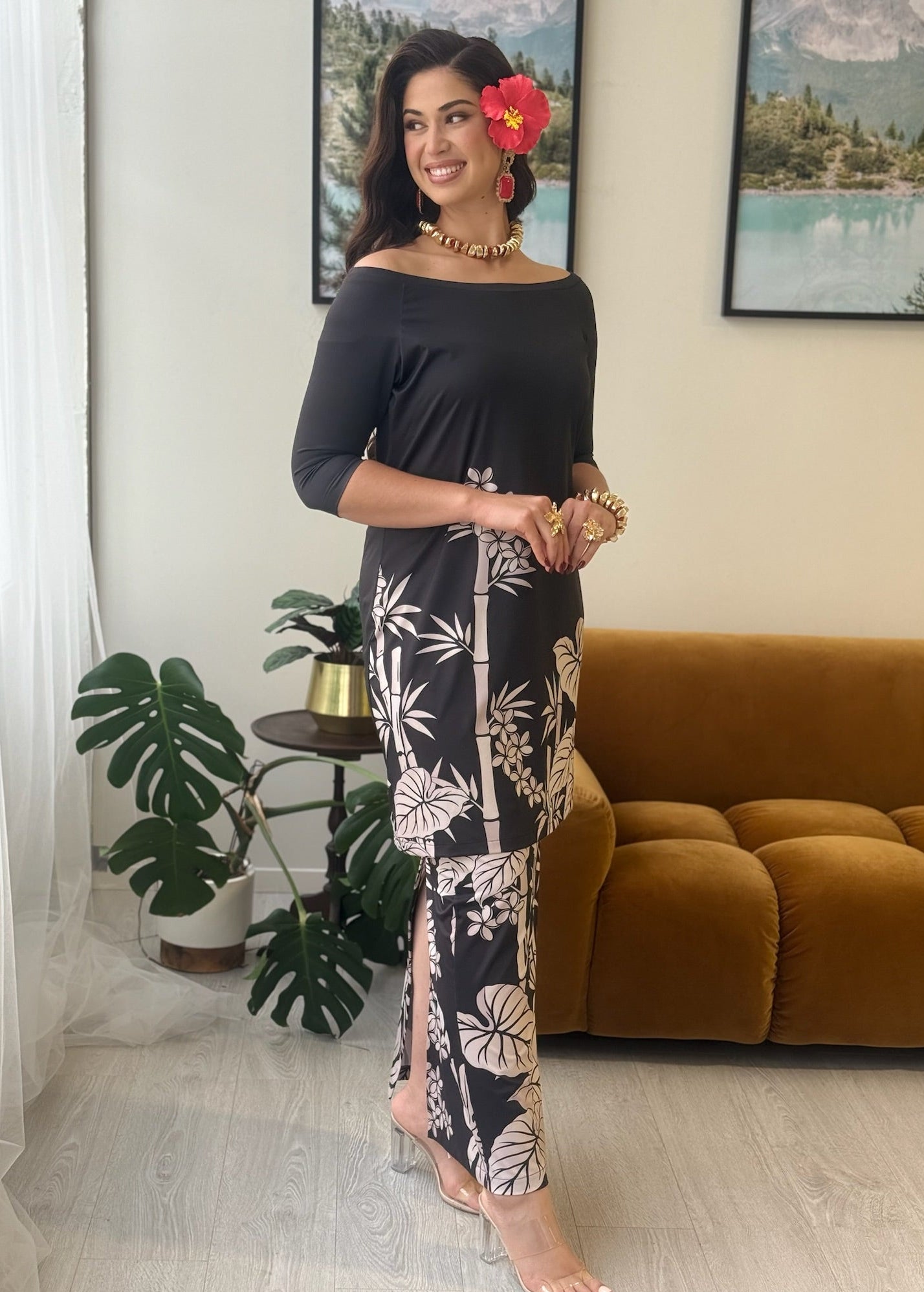 A woman models a black off-the-shoulder maxi dress with a white tropical leaf print. She is standing in a modern living room setting with a brown velvet sofa, potted plants, and framed landscape art on the wall.