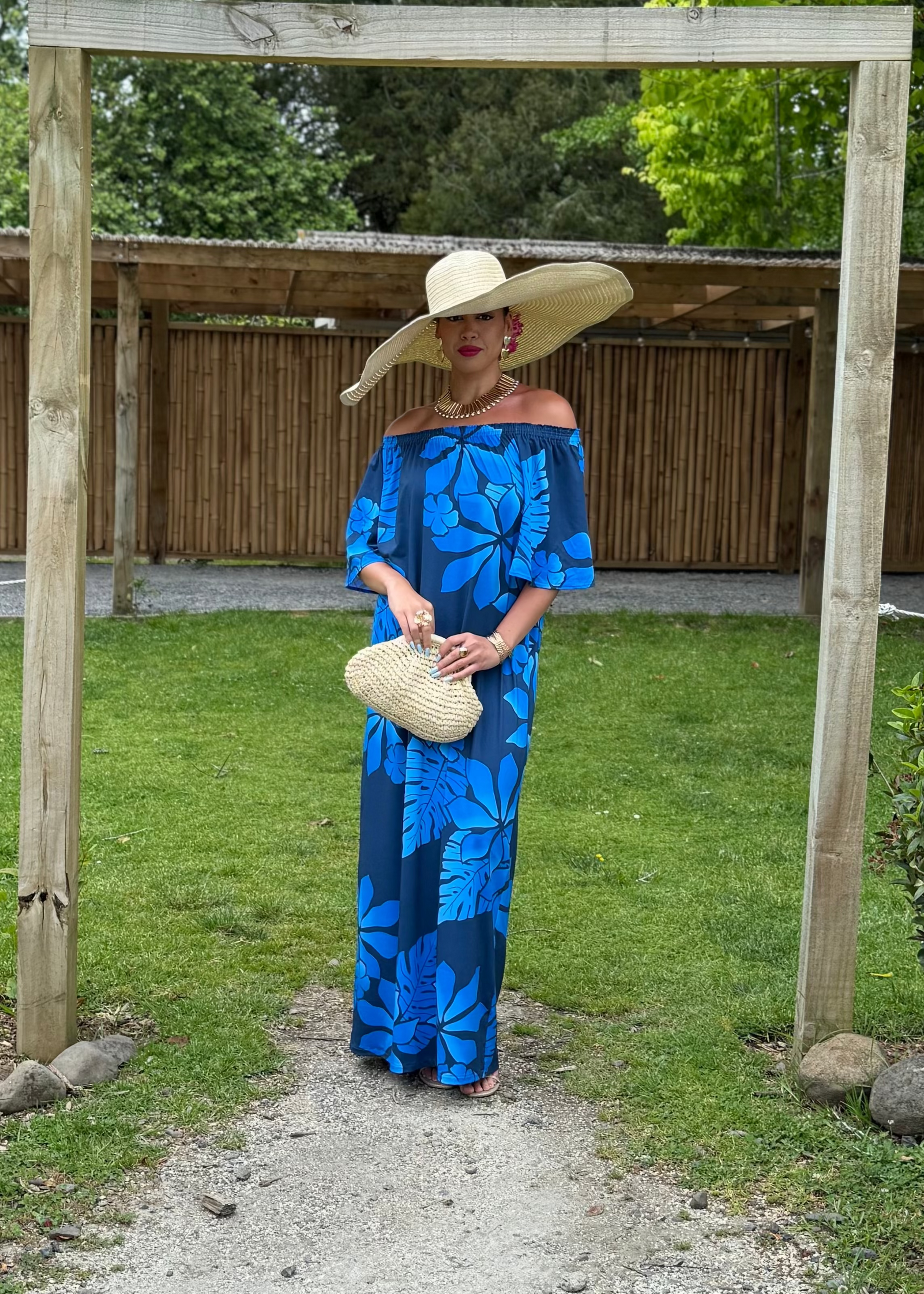 Woman in a blue dress and wide-brimmed hat standing under a wooden archway with greenery around.