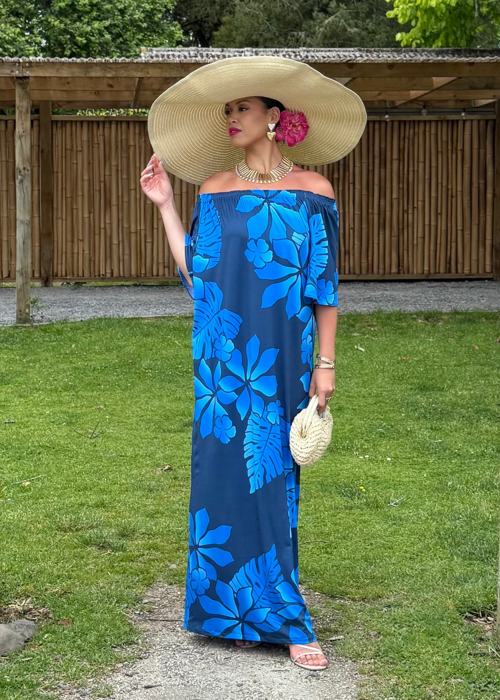 Woman in a blue floral dress and large hat standing under a wooden archway with greenery around.