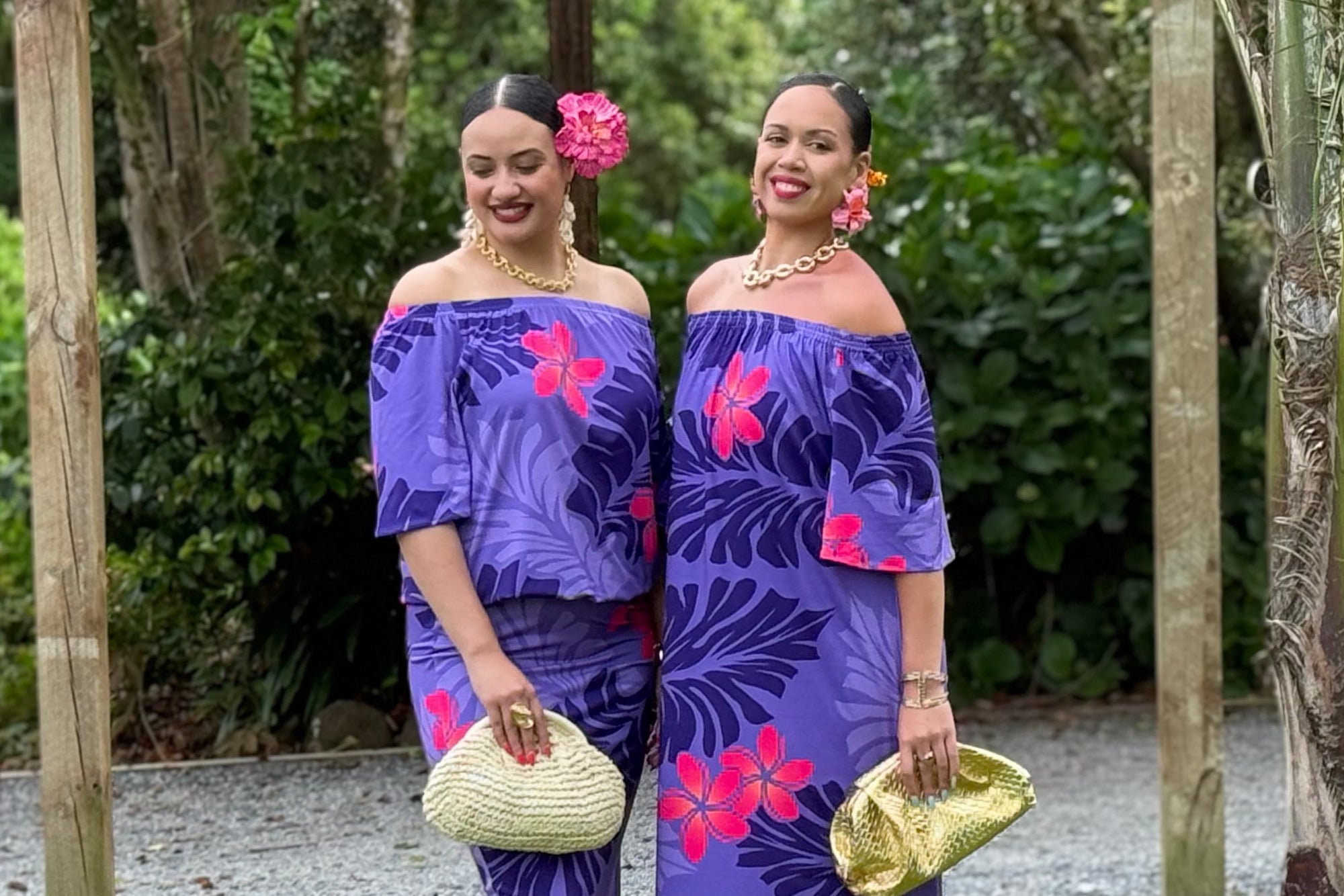 Two women in matching purple floral outfits standing outdoors with greenery in the background.