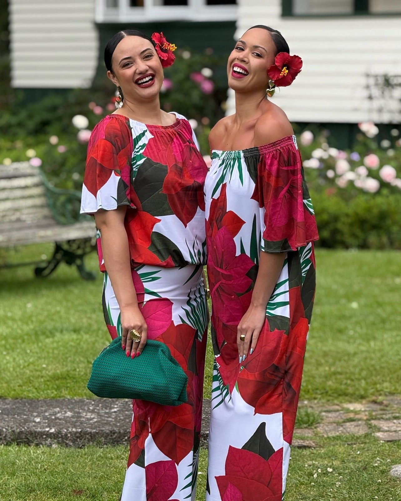Two women in colorful floral outfits standing outdoors with a house and garden in the background.