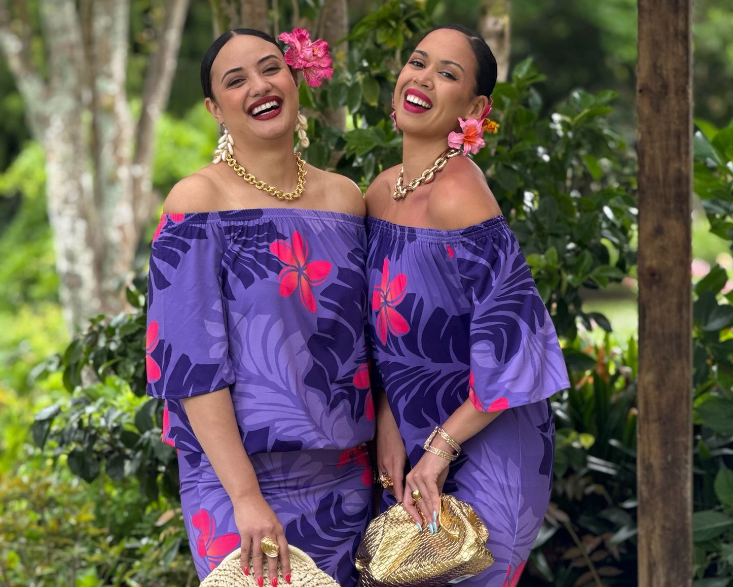 Two women in matching purple outfits with floral patterns standing in a lush green outdoor setting.
